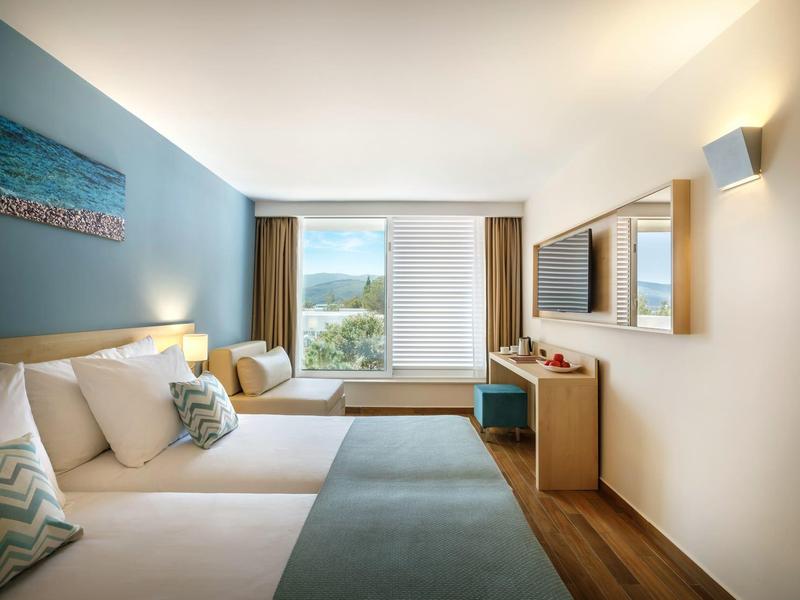 Modern hotel room with two beds, large window, and view of green hills.