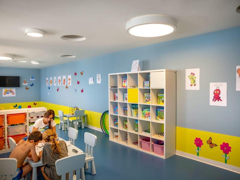 Colorful playroom with child tables, chairs, shelves, and a bulletin board on the blue wall.
