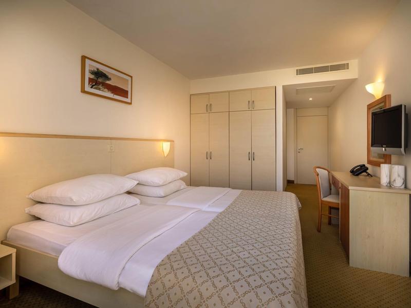 Helles Hotelzimmer mit großem Doppelbett, Schrank, Schreibtisch und TV an der Wand.