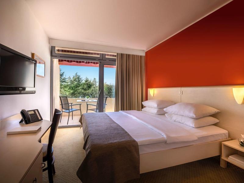 Modernes Hotelzimmer mit Doppelbett, roter Akzentwand, Balkon und hellem Parkettboden.