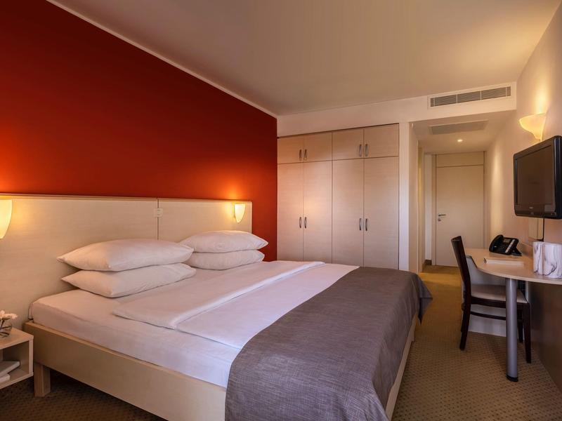 Modernes Hotelzimmer mit großem Bett, roter Wand, Schreibtisch und Wandfernseher.