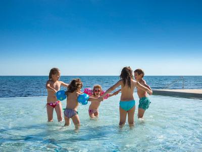 Fünf Kinder spielen lachend im flachen Wasser eines Pools mit klarem blauen Himmel.