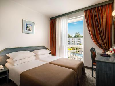 Kleines, helles Hotelzimmer mit zwei Betten, Balkon und braunen Vorhängen.