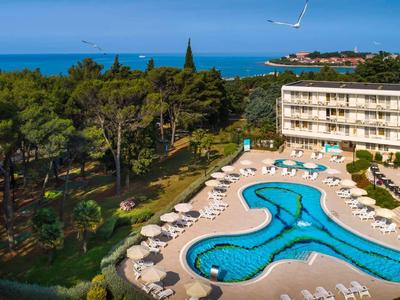 Piscina dell'hotel con lettini circondata da alberi e vista sul mare sotto un cielo limpido.