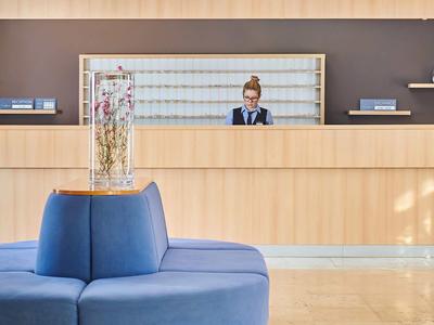 Banco reception in legno chiaro con divano blu davanti e vaso con fiori secchi.