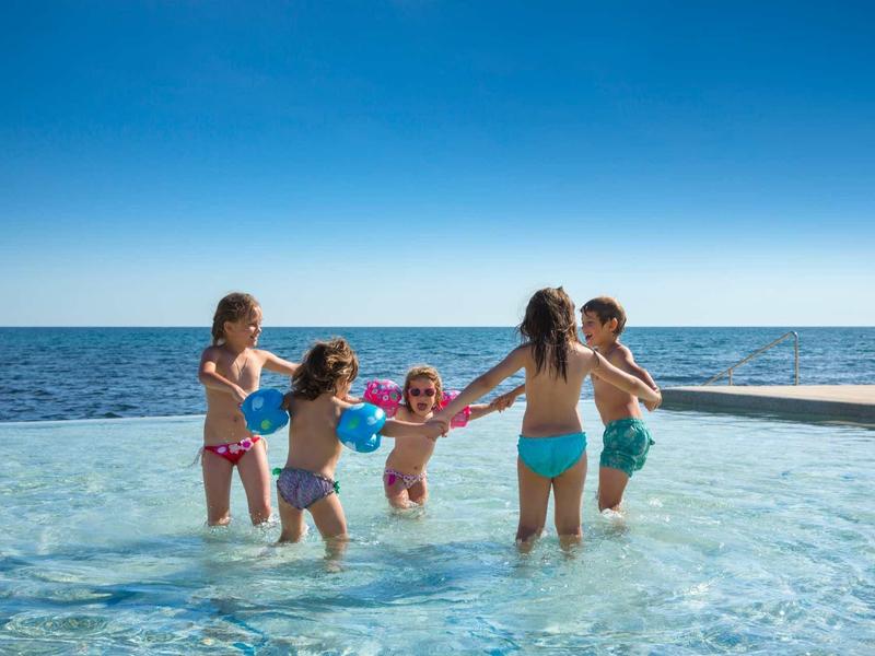 Fünf Kinder spielen lachend im flachen Wasser eines Pools mit klarem blauen Himmel.
