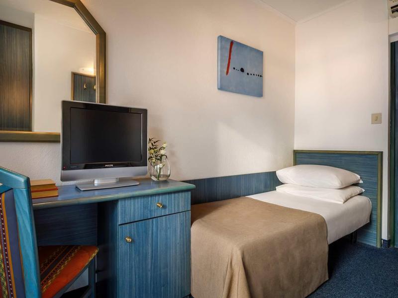 Kleines Zimmer mit Einzelbett, blauem Schreibtisch, Stuhl und kleinem Fernseher an der Wand.