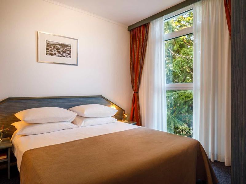 Gemütliches Schlafzimmer mit braunem Bett, zwei Lampen, Vorhängen und Fenster mit Blick ins Grüne.