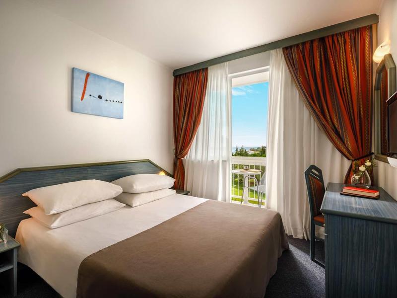 Gemütliches Hotelzimmer mit Doppelbett, brauner Bettdecke, roter Gardine und Schreibtisch am Fenster.
