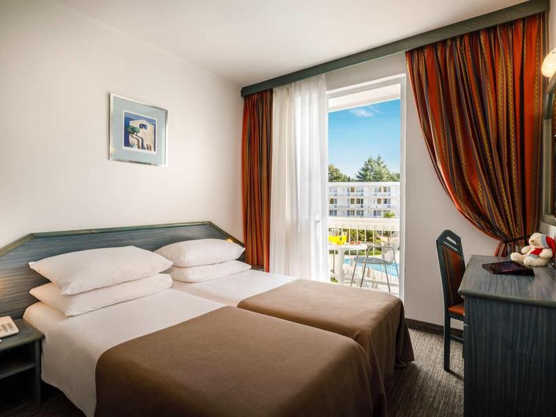 Kleines, helles Hotelzimmer mit zwei Betten, Balkon und braunen Vorhängen.