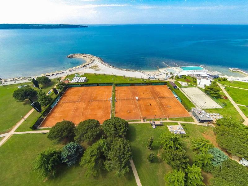 Luftaufnahme von zwei orangefarbenen Tennisplätzen am Meer, umgeben von grünem Gras und Bäumen.