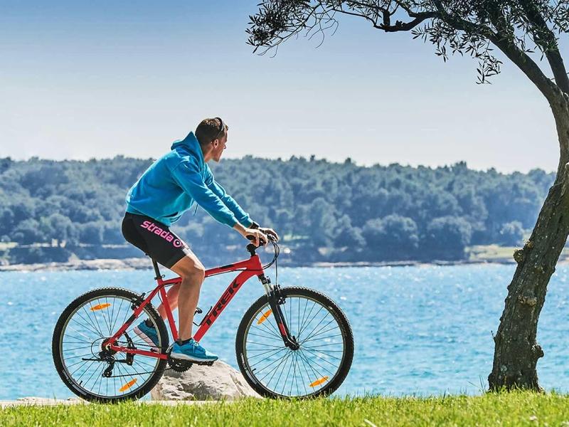 Ein Mann fährt mit einem roten Fahrrad am Wasser entlang bei sonnigem Himmel und grünem Gras.