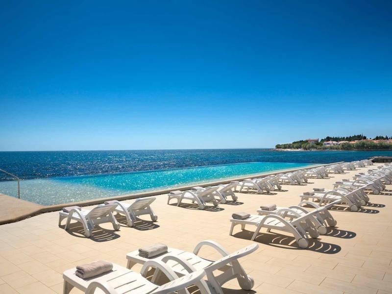 Meerblick mit Infinity-Pool und weißen Sonnenliegen an sonnigem Tag unter blauem Himmel.