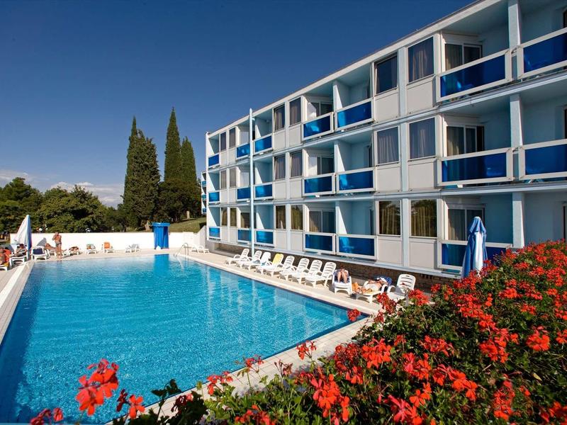Großes Außenbecken neben modernem Hotelgebäude mit blauen Balkonen, Sonnenliegen und blühenden Sträuchern.