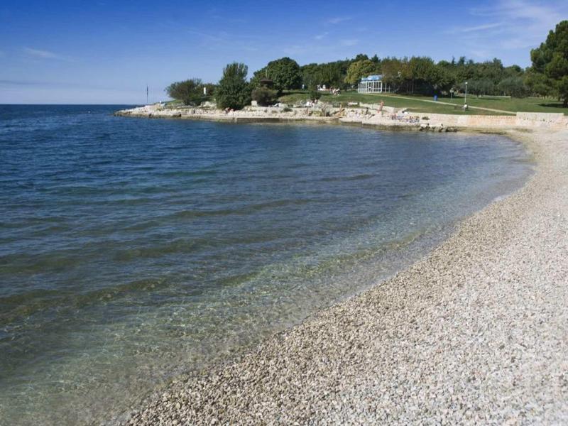 Kieselstrand mit klarem Wasser und bewaldetem Ufer unter blauem Himmel.