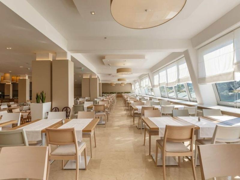 Heller, großer Speisesaal mit Holzstühlen und Tischen, großen Fenstern und Deckenlampen.