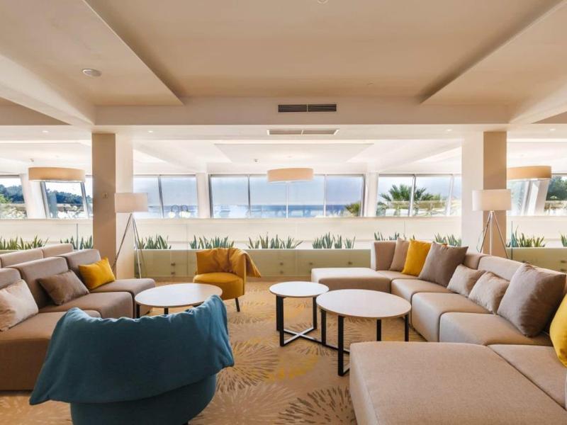 Helle Lounge mit großen Fenstern, gemütlichen Sofas in Beige und Senfgelb, runden Tischen und weichen Teppichen.