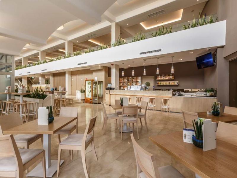 Moderner Cafébereich mit Holzstühlen, Tischen und hellen Wänden, großer Bar und Pflanzen.