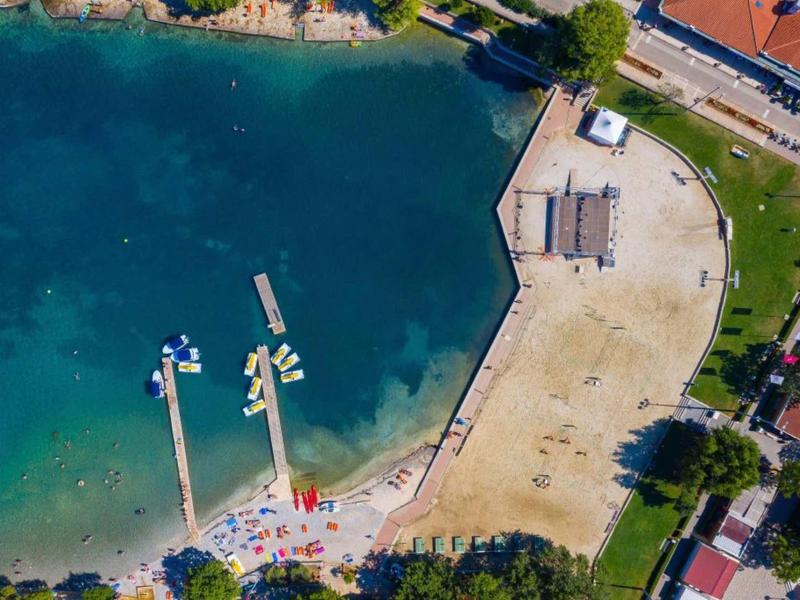 Luftaufnahme eines Strandes mit klarem blauen Wasser, zwei langen Stegen und angrenzendem Sandbereich.