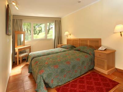 Habitación de hotel luminosa con dos camas individuales, muebles de madera y ventana con vistas al verde.