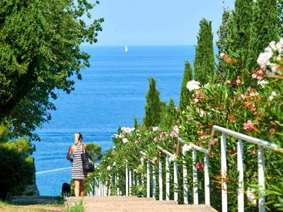 Vrouw loopt op een pad met groen en bloemen naar de zee met een zeilboot op de achtergrond.
