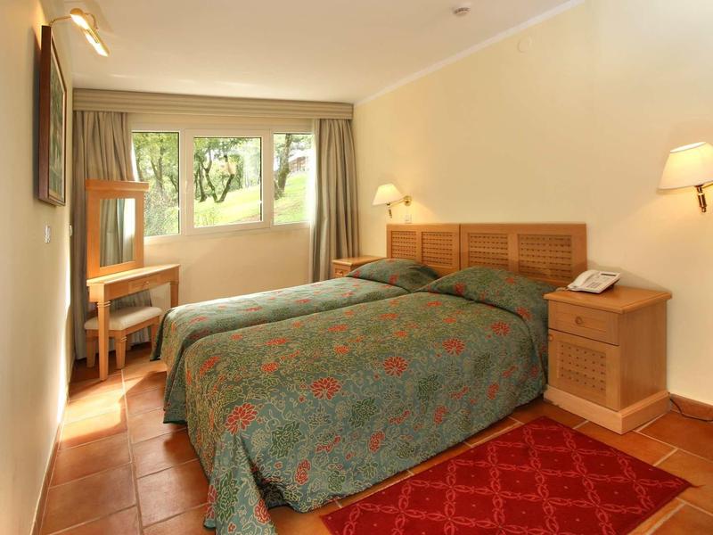 Habitación de hotel luminosa con dos camas individuales, muebles de madera y ventana con vistas al verde.