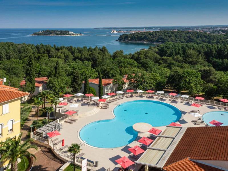 Vista de una piscina de hotel con sombrillas rojas, rodeada de vegetación y mar al fondo.