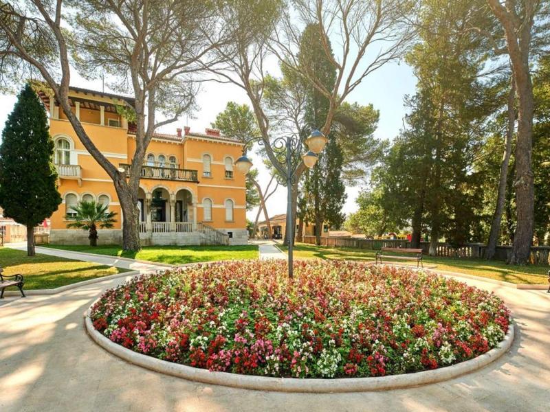 Edificio histórico de hotel detrás de un parterre circular de flores en un parque soleado con árboles altos.