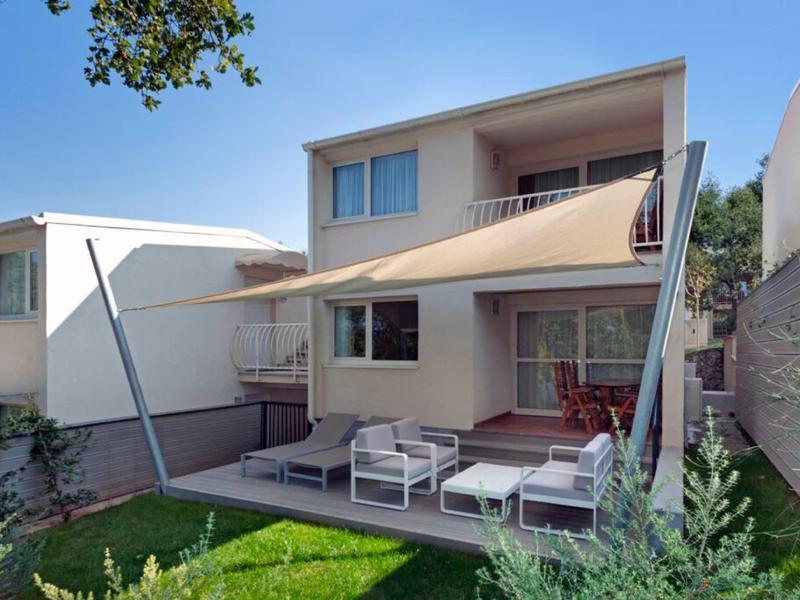 Jardín moderno con terraza, toldo y área de descanso frente a una casa de dos pisos.