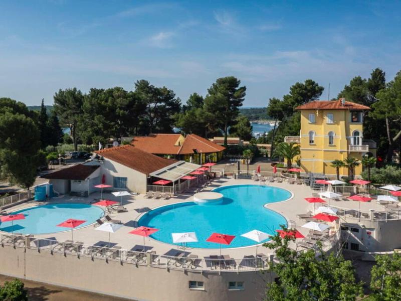 Piscina redonda de hotel con sombrillas rojas y edificio amarillo al fondo en un día soleado.