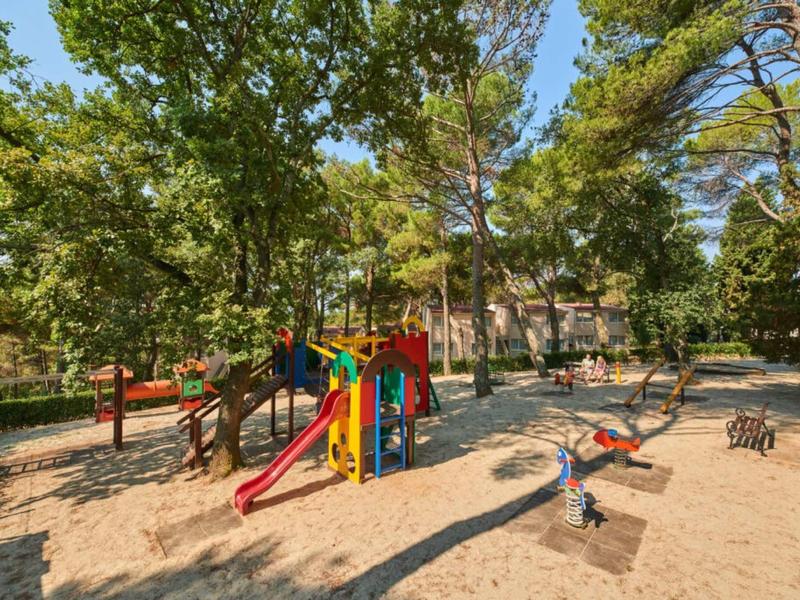 Parque infantil con tobogán colorido y balancín rodeado de árboles en un parque soleado.