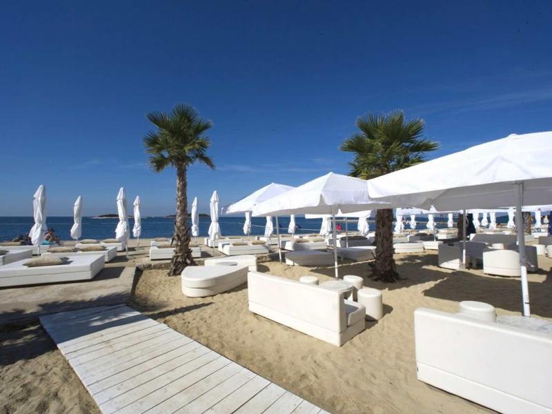 Lounge de playa con tumbonas blancas, sombrillas y palmeras bajo un cielo azul despejado.