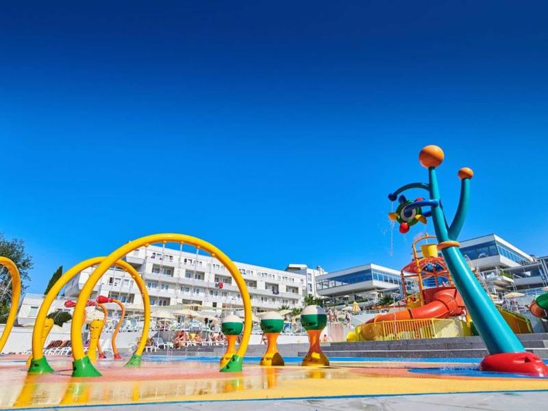 Parque acuático colorido con toboganes y fuentes frente a un hotel bajo cielo azul.