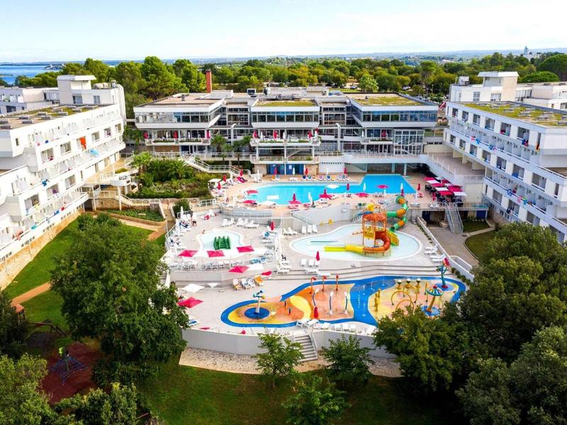 Resort con varias piscinas, áreas de juegos y edificios blancos rodeados de vegetación y mar al fondo.