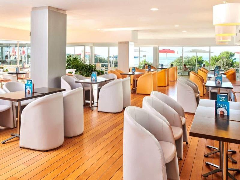 Área de lounge de hotel luminosa con suelo de madera, sillas cómodas y grandes ventanas con vista.