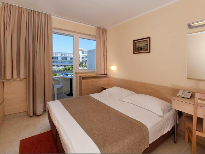 Habitación de hotel sencilla con cama doble, escritorio y balcón con vista a otros edificios.