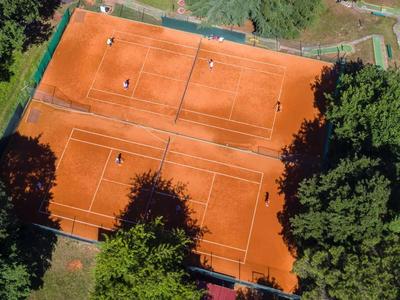Vier rote Sandplatz-Tennisplätze umgeben von Bäumen und Grünflächen.