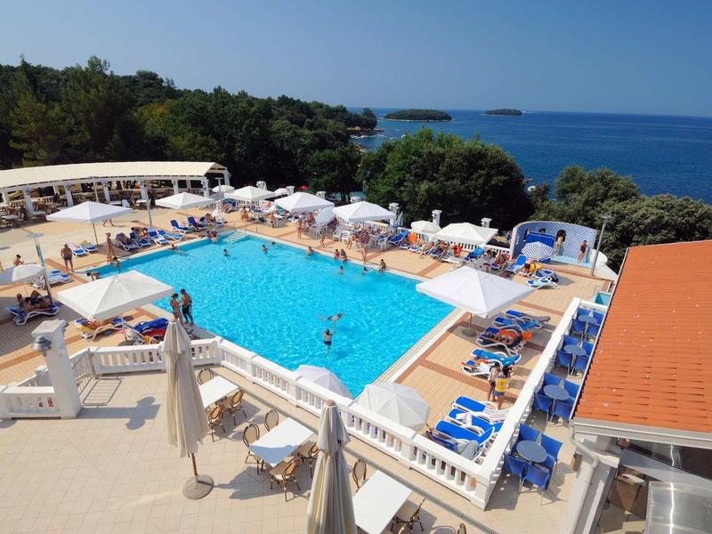 Piscina dell'hotel luminosa con lettini, ombrelloni e vista sul mare circondata da alberi.