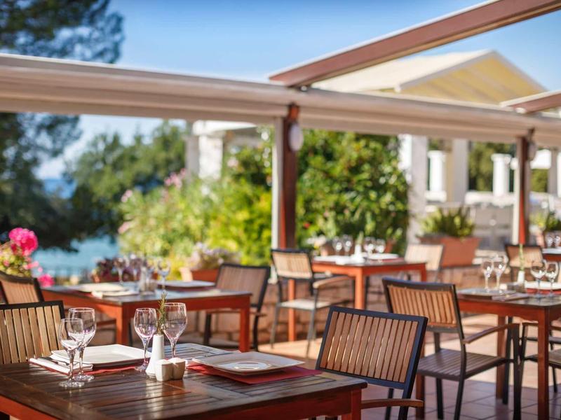 Terrazza con tavoli apparecchiati e sedie, circondata da vegetazione lussureggiante e vista sull'acqua.