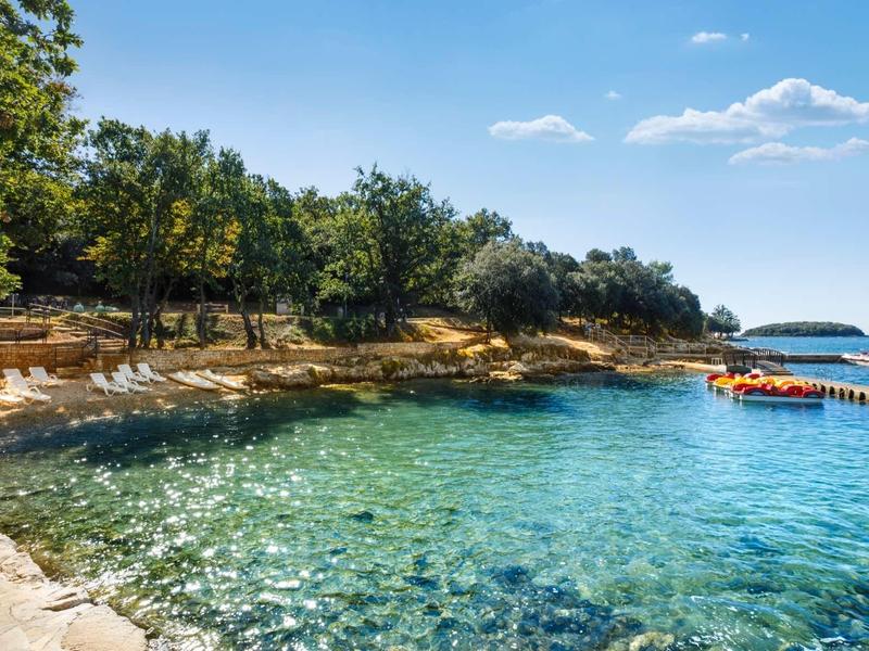 Acqua turchese limpida lungo costa rocciosa con alberi e piccolo molo sotto cielo soleggiato.