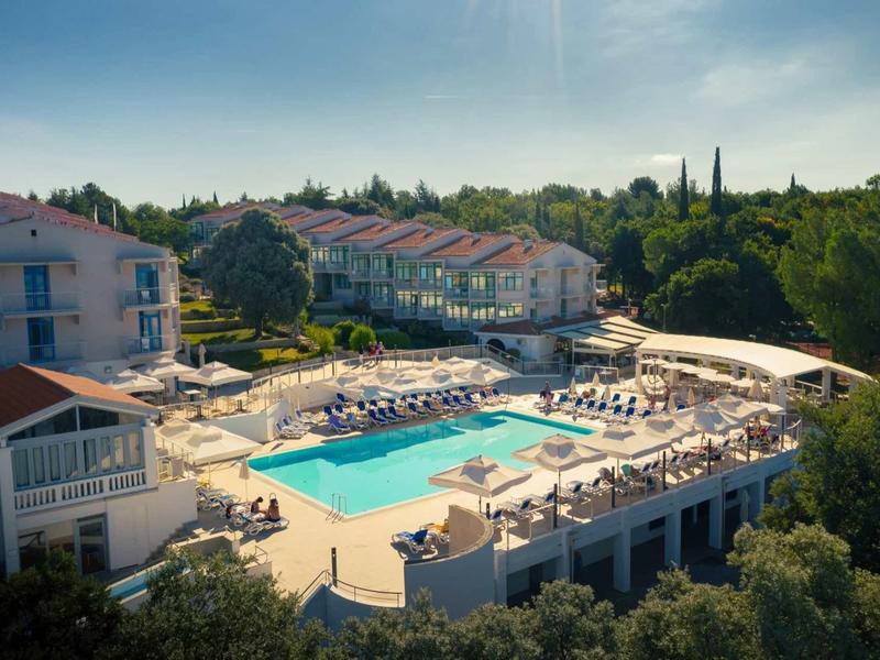 Complesso alberghiero con grande piscina, lettini e ambiente verde sotto un cielo sereno.