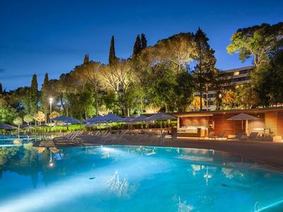 Oświetlony basen hotelowy nocą otoczony drzewami i budynkami.