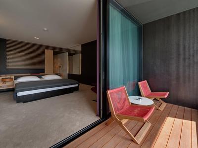 Modernes Hotelzimmer mit großem Bett und Balkon mit zwei roten Stühlen und Holzboden.
