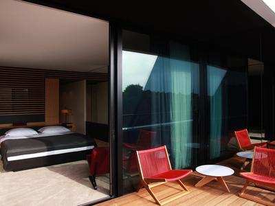 Balkon mit roten Stühlen und weißem Tisch vor einem Hotelzimmer mit Doppelbett.
