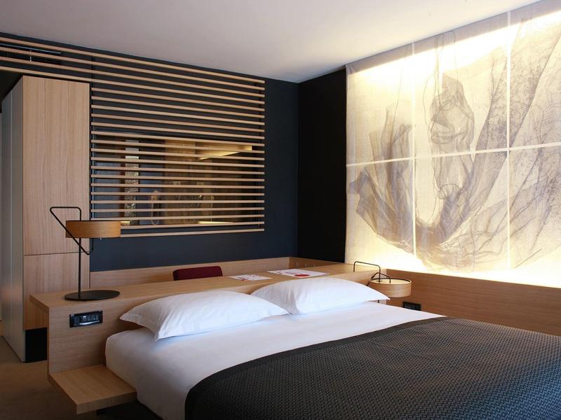 Nowoczesny pokój hotelowy z dużym łóżkiem, stylowym oświetleniem ściennym i minimalistycznym umeblowaniem.