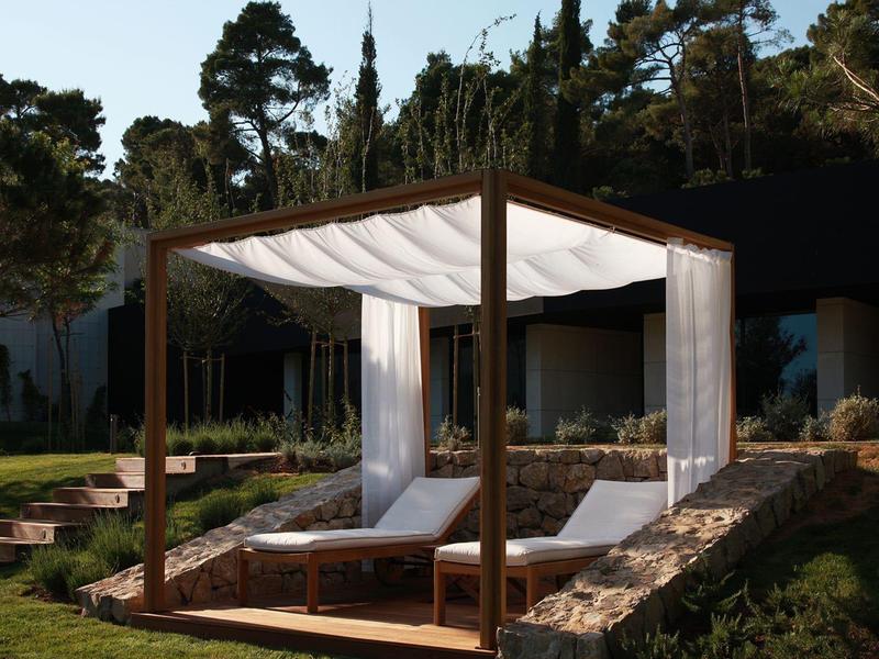 Drewniany pawilon z białymi zasłonami nad dwoma leżakami w ogrodzie.