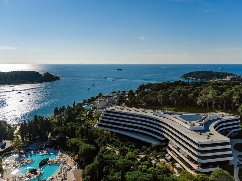 Hotel o zakrzywionym kształcie nad morzem z dużym basenem w słoneczny dzień.