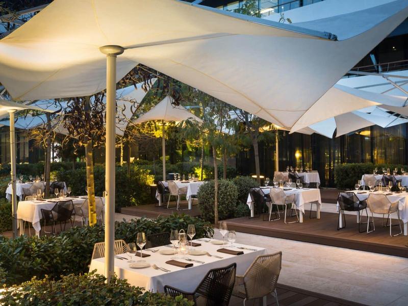 Elegancka restauracja na zewnątrz z nakrytymi stołami pod białymi żaglami oświetlona wieczornym światłem.