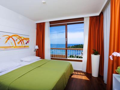 Hell eingerichtetes Hotelzimmer mit großem Bett, grünem Bettbezug und Balkon mit Meerblick.