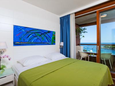 Helles Hotelzimmer mit grünem Bett, blauem Vorhang und Meerblick durch große Fenster.